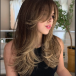 A person with long, layered brown hair and bangs is looking to the side in a modern indoor setting, wearing a black sleeveless top.